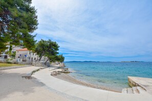 A coastal walkway with steps leading to the sea, surrounded by trees and buildings with a clear view of the water and distant islands.