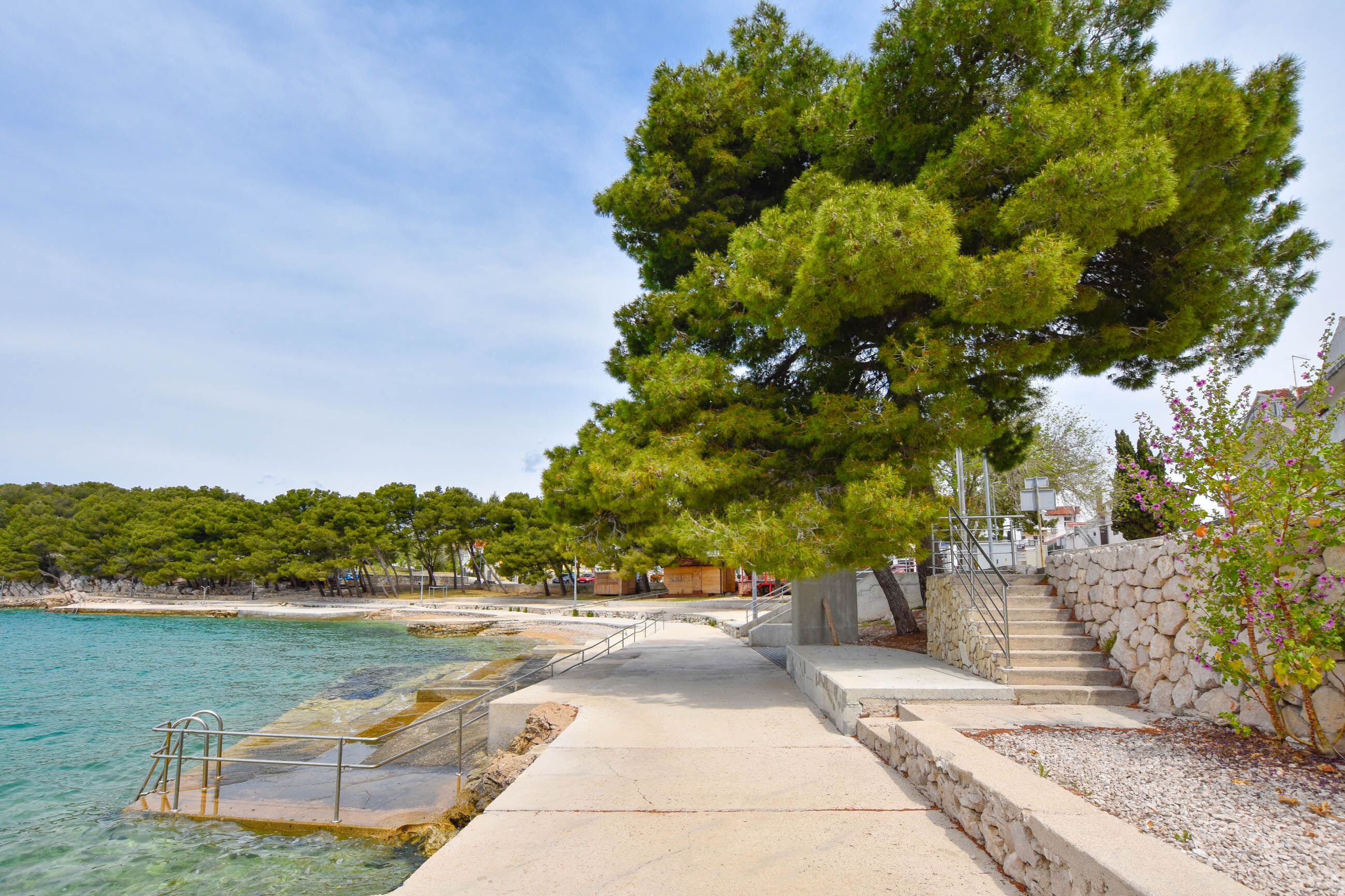 Une promenade en bord de mer menant à une plage à l’eau claire, avec un grand arbre et un escalier en pierre.