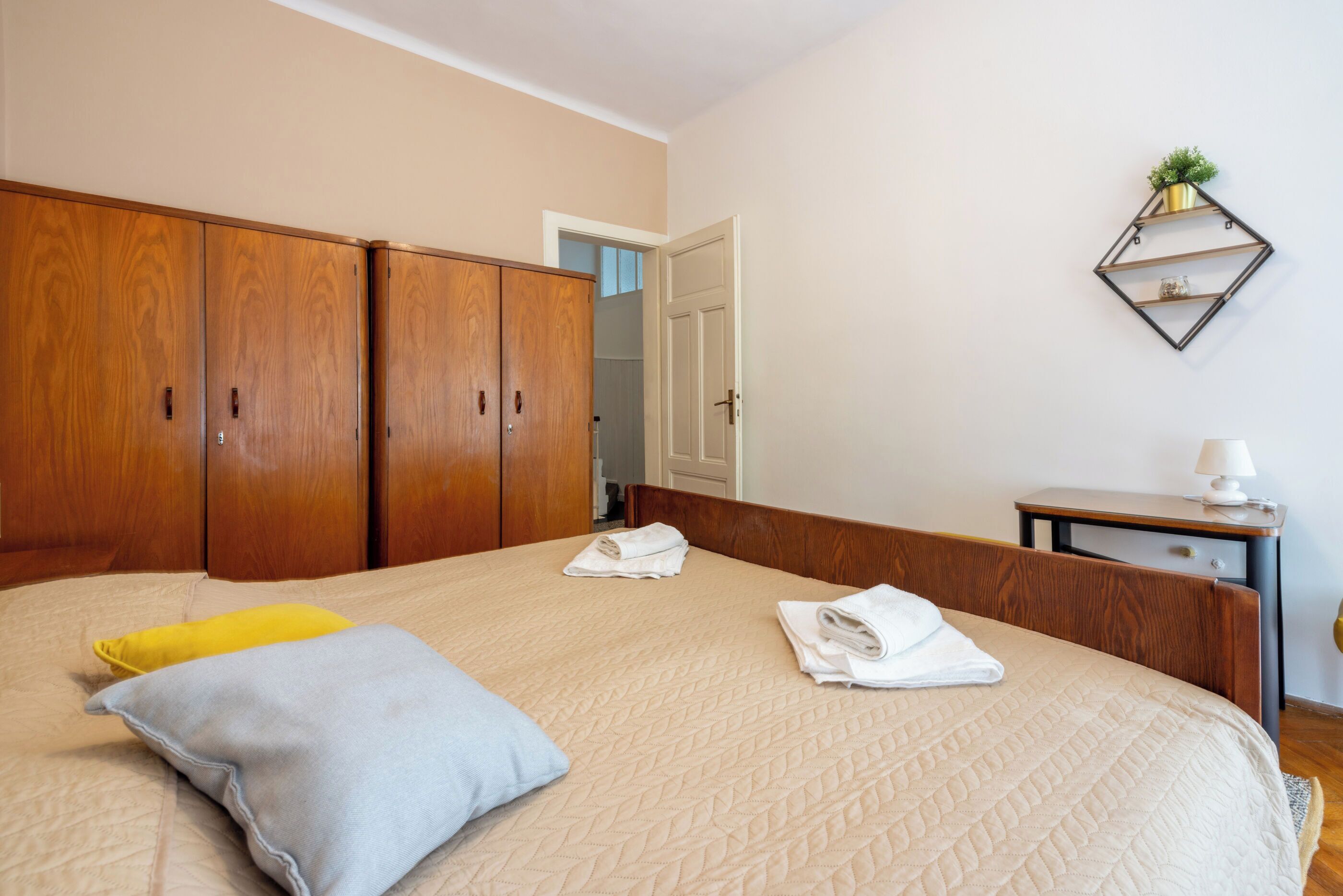 Un dormitorio con una cama, un armario de madera, una mesita de noche con una lámpara y una estantería en la pared con una planta.