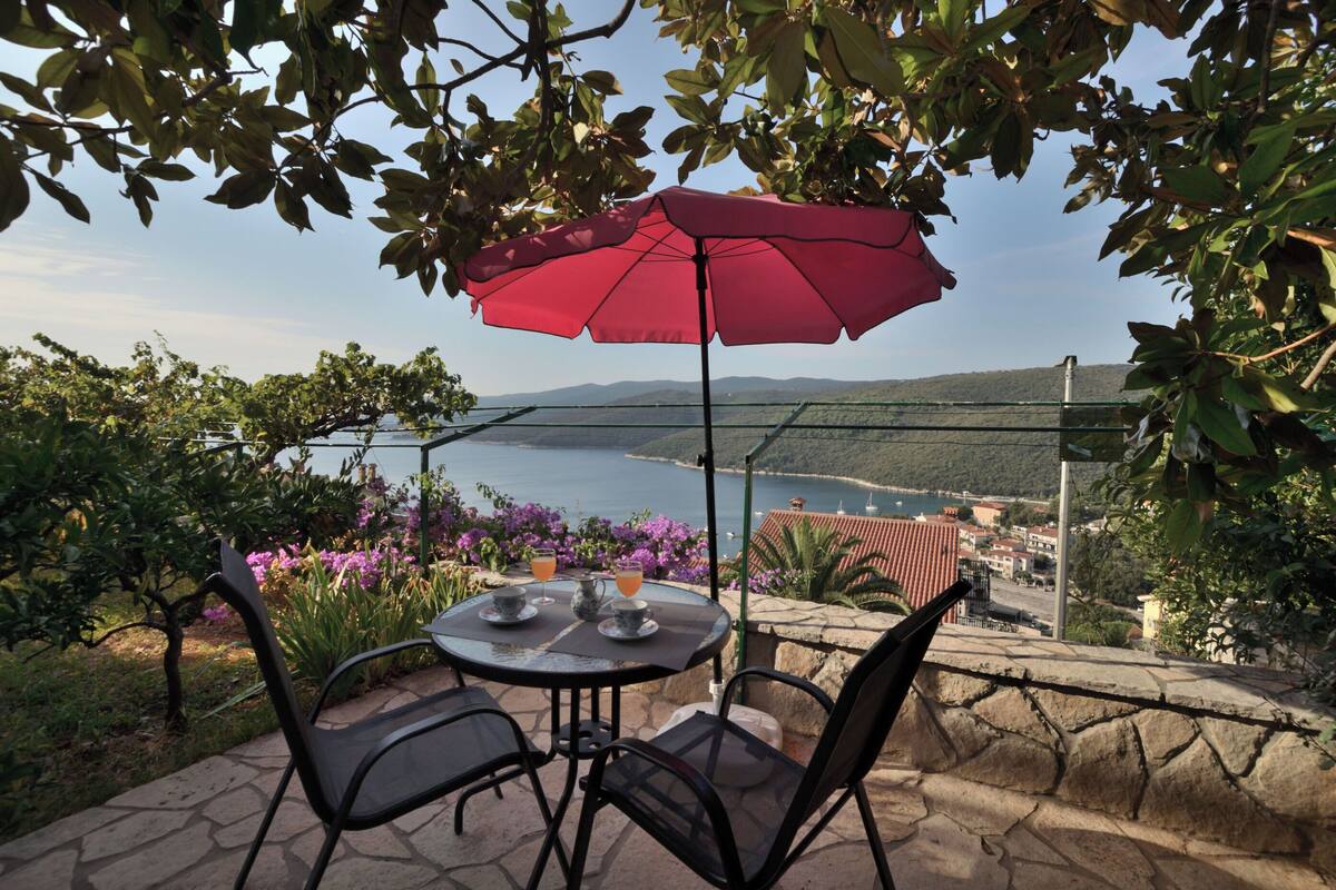 A table with chairs and a red umbrella, overlooking a scenic view of a body of water and hills.