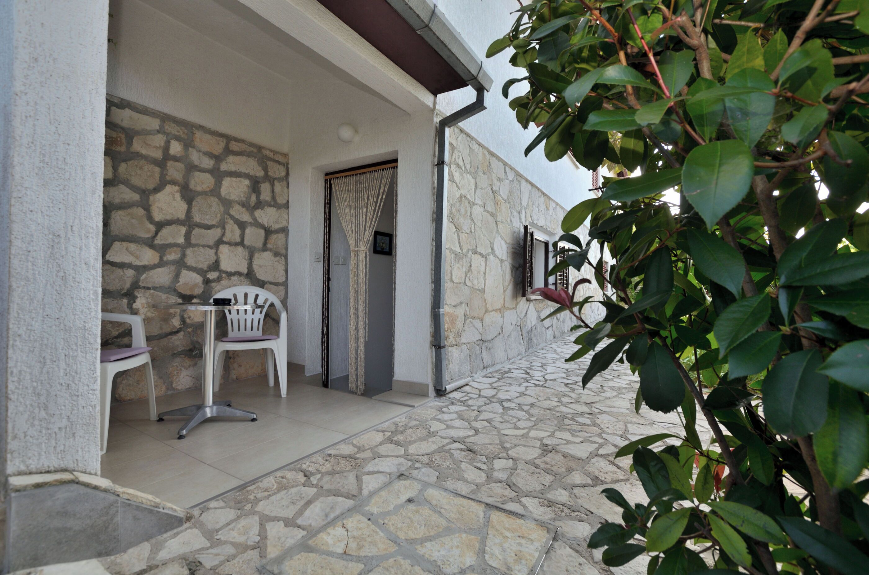 Eine Terrasse mit Steinboden, zwei weiße Stühle, ein Tisch, eine Steinmauer und ein Fenster mit Vorhang.