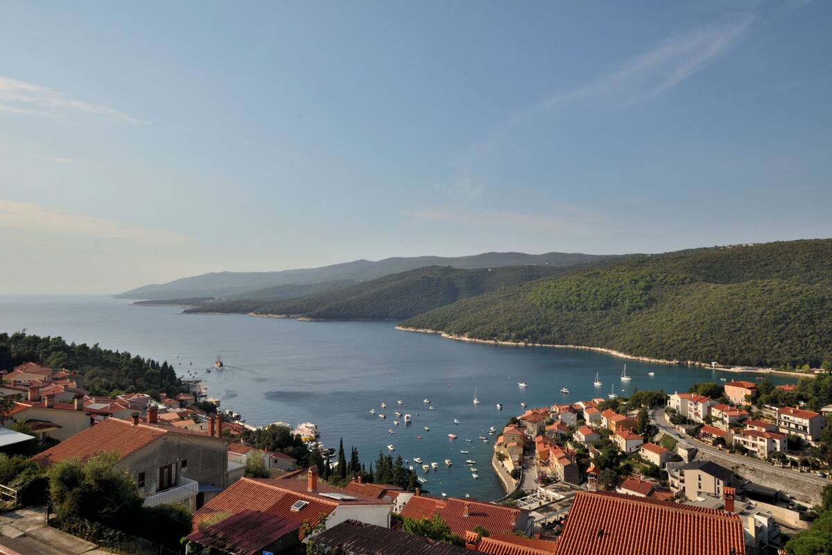 A coastal town with red-roofed buildings, a marina with boats, and a clear view of the sea and distant hills.