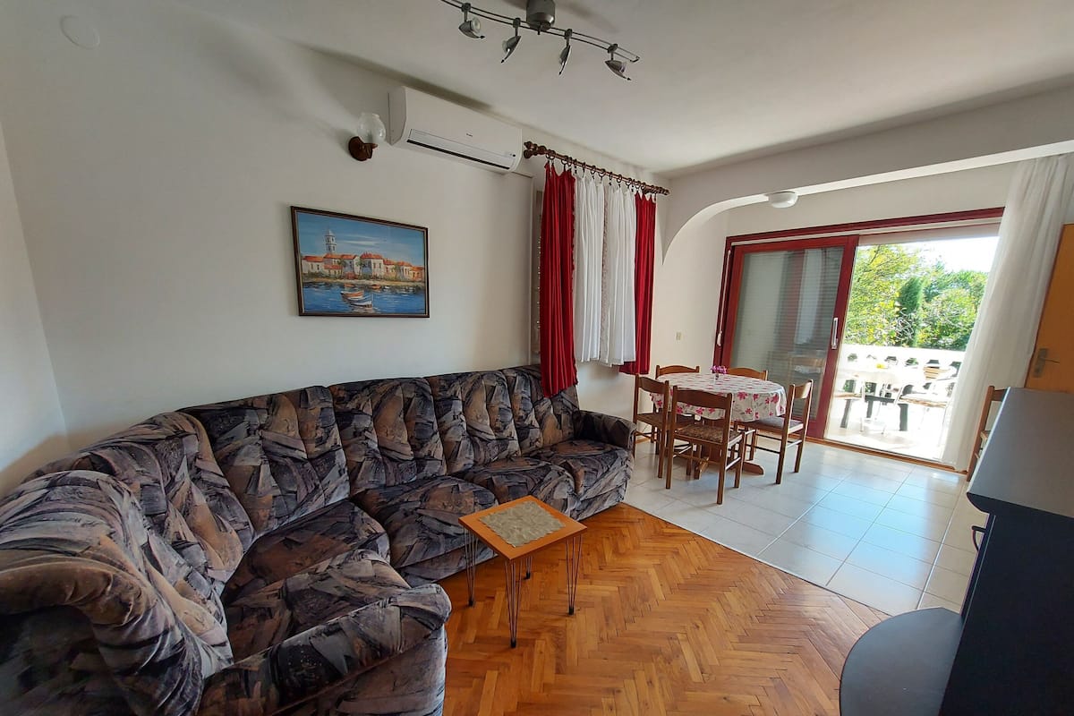 Un salón con un sofá estampado, una mesita, una zona de comedor con sillas, un cuadro en la pared y un ventanal corredero que da a un espacio exterior.