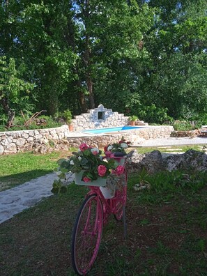 Ein rosafarbenes Fahrrad mit Blumenkorb, eine Steinmauer und ein kleiner Pool in einem Garten.
