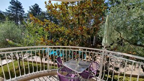 Un balcone con tavolo in vetro e sedie viola, circondato da una ringhiera bianca e vegetazione.