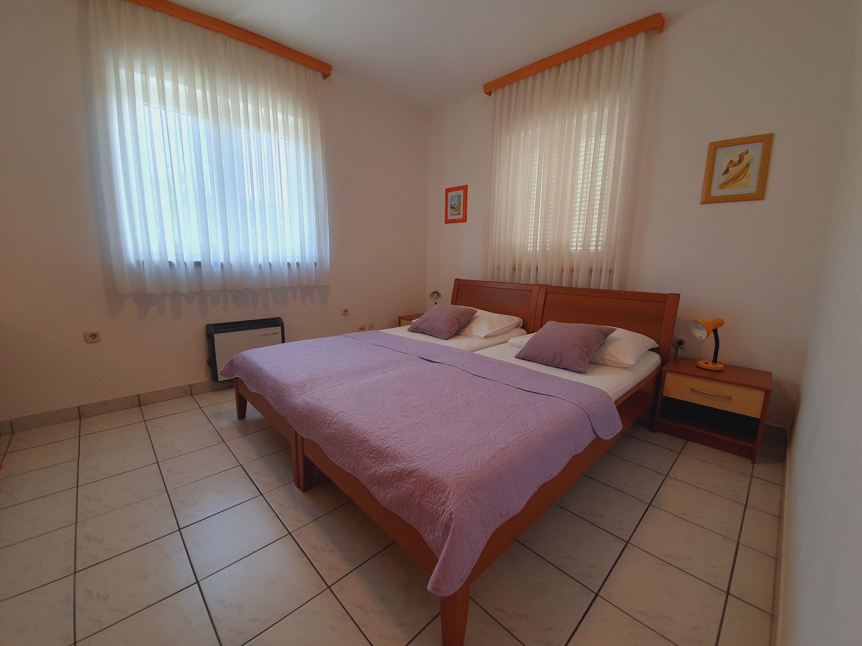 Un dormitorio con una cama grande, dos mesitas de noche, un calentador y una ventana con cortinas traslúcidas.