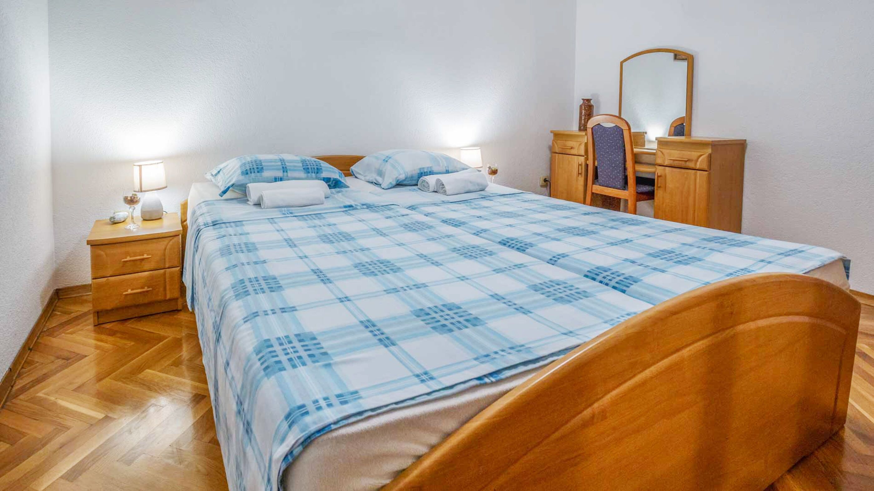 Un lit bien fait, recouvert d’une couverture à carreaux bleus et blancs, une table de chevet en bois avec une lampe, une commode avec un miroir et une chaise.
