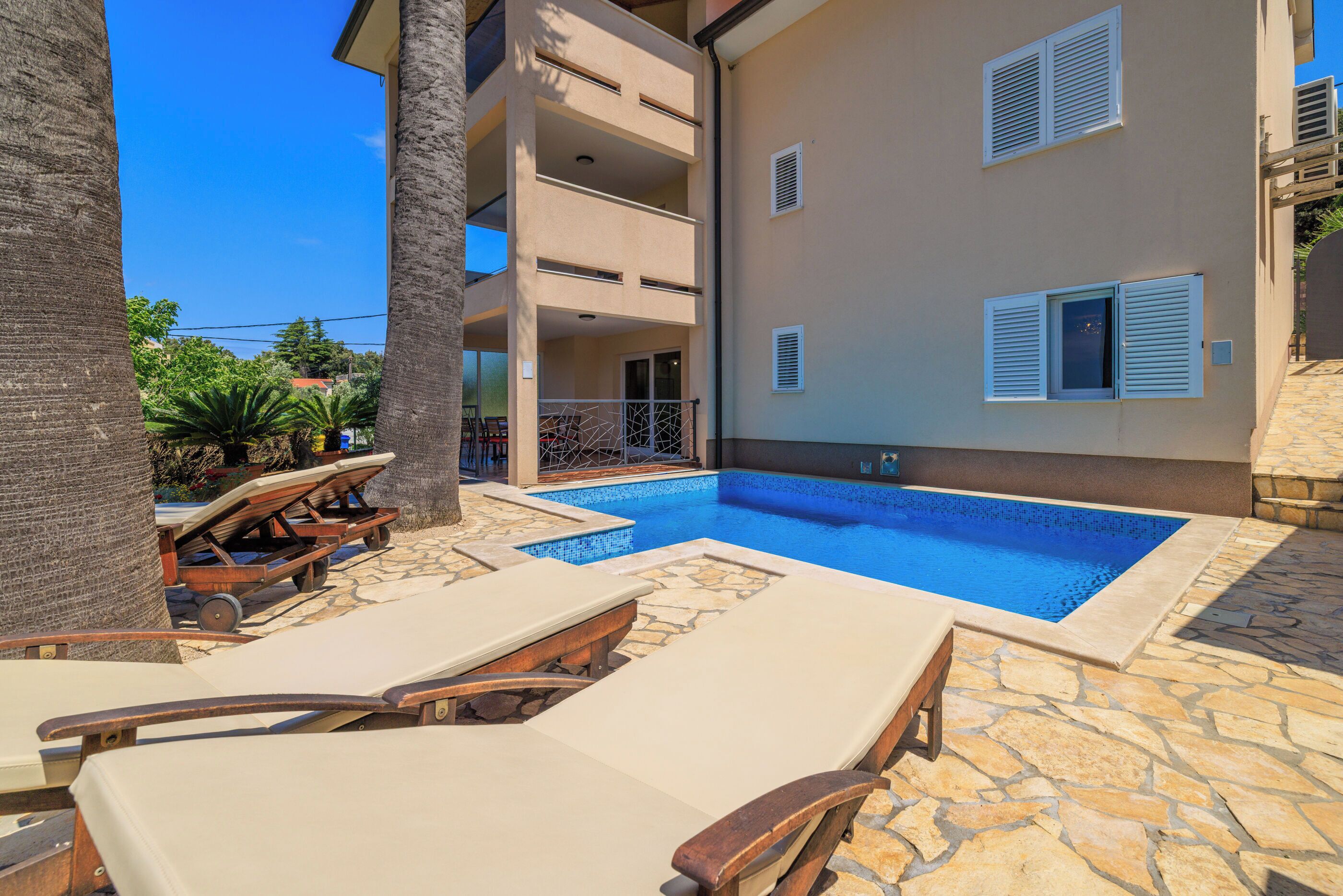 A pool area with lounge chairs, a stone-tiled deck, and a building with balconies and windows.
