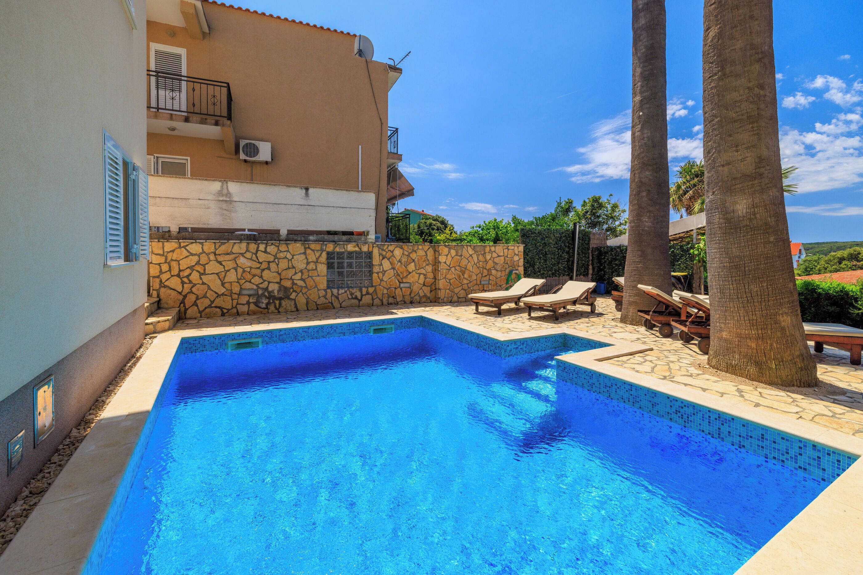 Une piscine avec des chaises longues, un mur en pierre et un bâtiment doté de balcons.