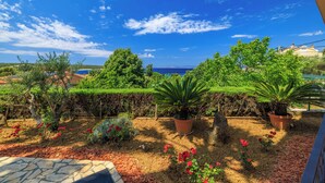 Ein Garten mit verschiedenen Pflanzen und Blumen, einem Kiesweg und Blick auf das Meer sowie Gebäude im Hintergrund.