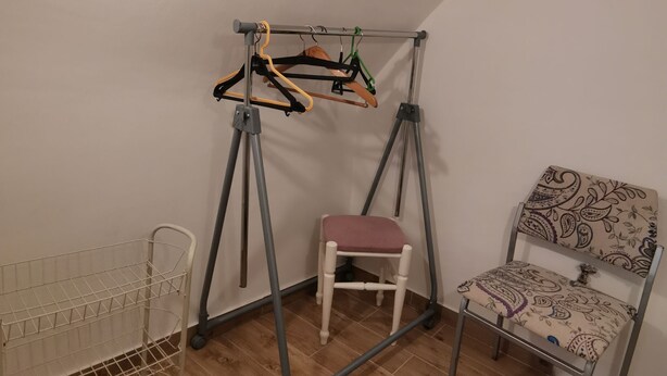 A room with a wooden floor, a metal chair with a patterned cushion, a metal shelf unit, and a metal swing set with clothes hangers. - Apartment Maja (Bribir)