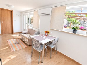 A modern living room with a sofa, a dining table with chairs, a small round table with a vase, and a window with plants.
