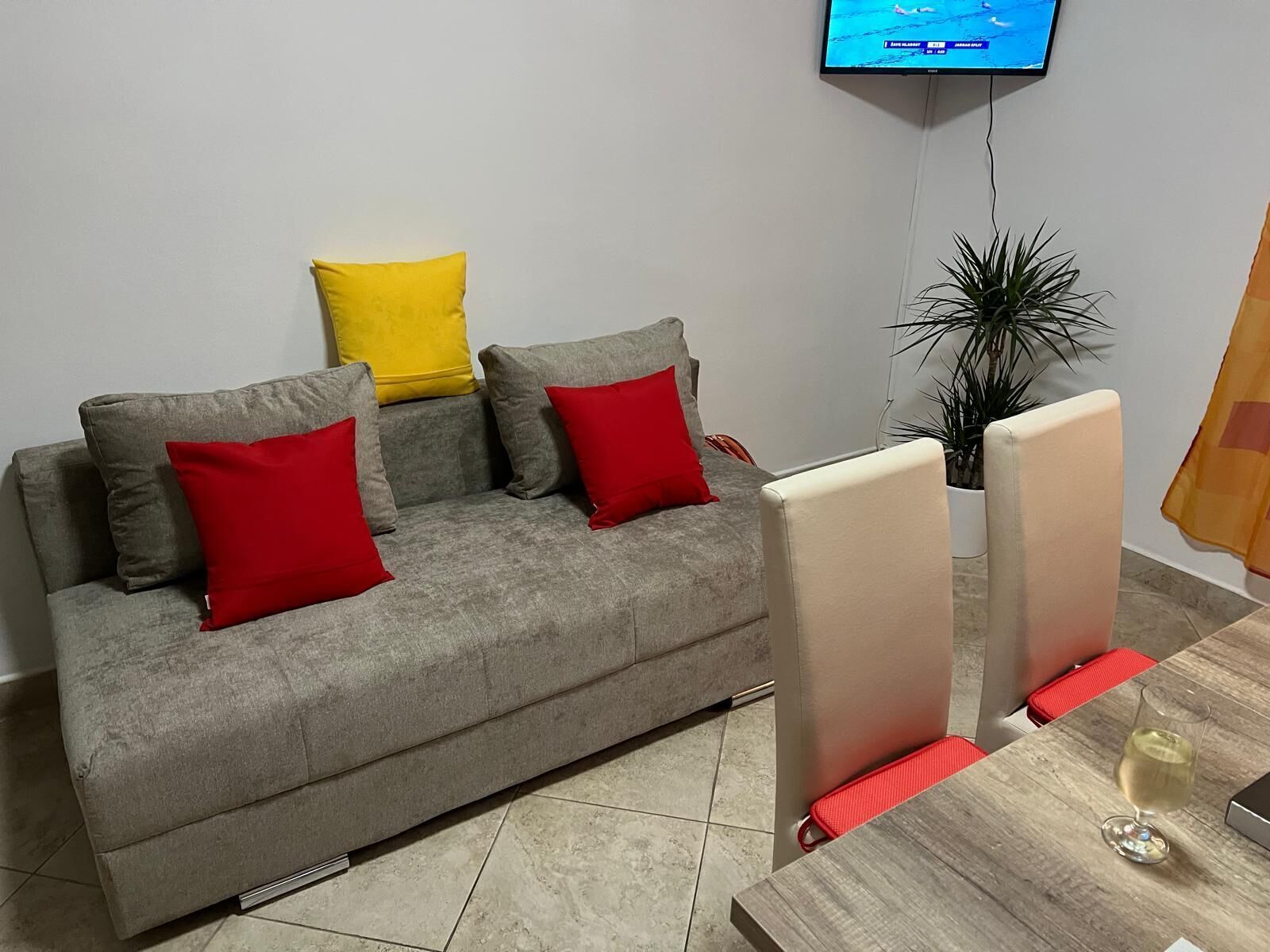 Un salón con un sofá gris, cojines rojos y amarillos, un televisor montado en la pared, una planta en maceta y una mesa de madera con una copa de vino.