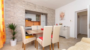 A modern kitchen with white cabinets, a wooden dining table, and chairs. There is a potted plant and a painting on the wall. - Apartments Milica (Kastel Stari)