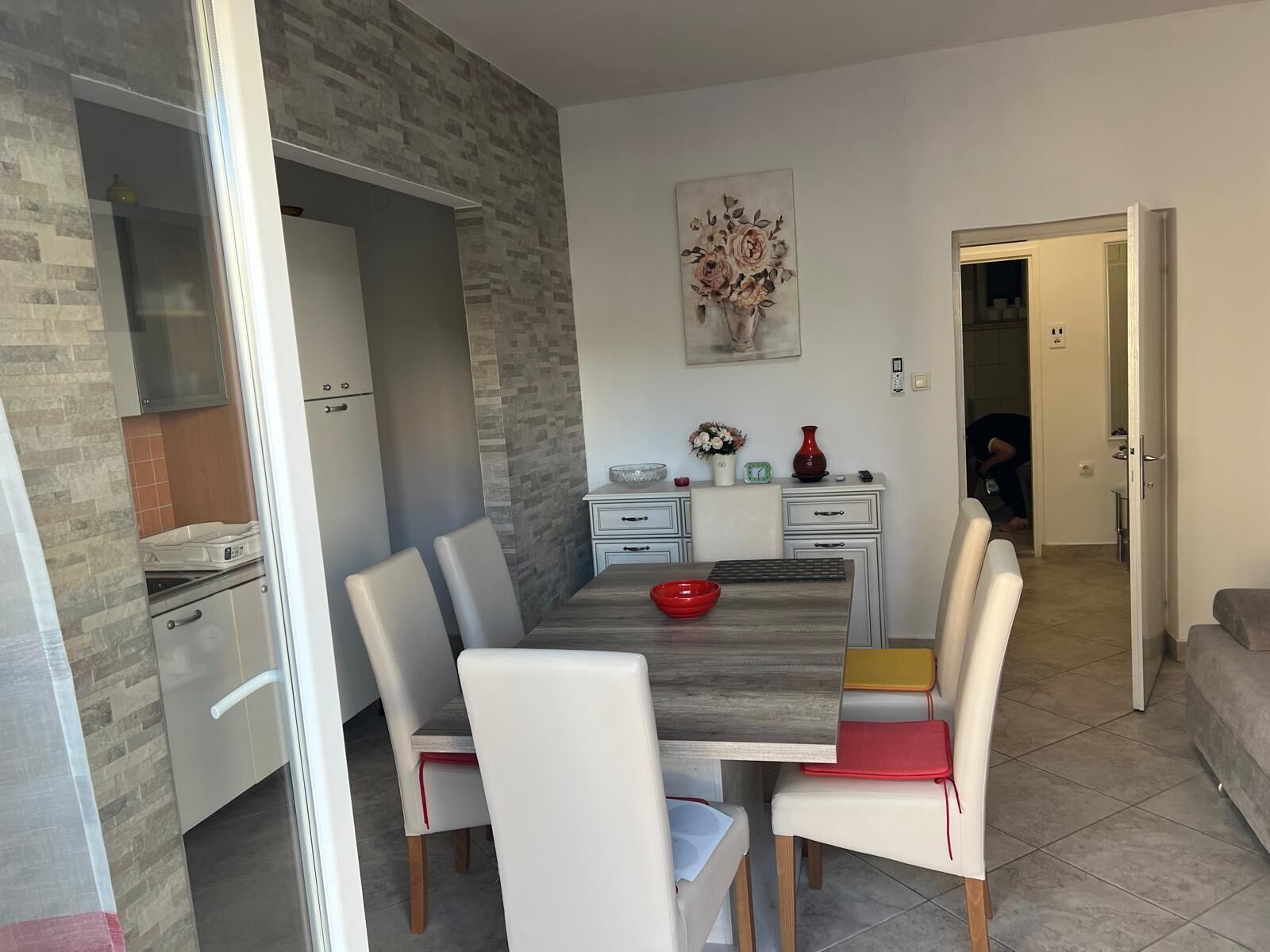 Un comedor con una mesa de madera, sillas blancas y un tazón rojo. Hay un armario blanco con cajones y un jarrón con flores encima. Un cuadro de flores cuelga en la pared.