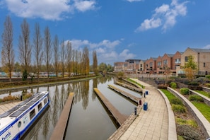 A canal with a boat docked on the left, a pedestrian walkway on the right, and a row of brick buildings in the background.