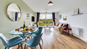 A modern living room with a glass dining table, blue chairs, a sofa, and a large window offering a view of the outdoors.