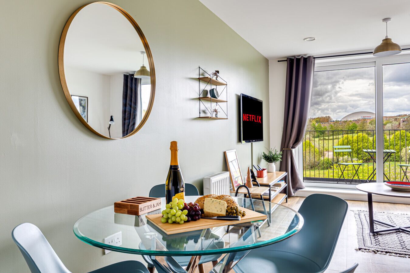 A modern living room with a glass dining table, a bottle of champagne, and a fruit platter. A flat-screen TV is mounted on the wall, and a large window offers a view of the outdoors.