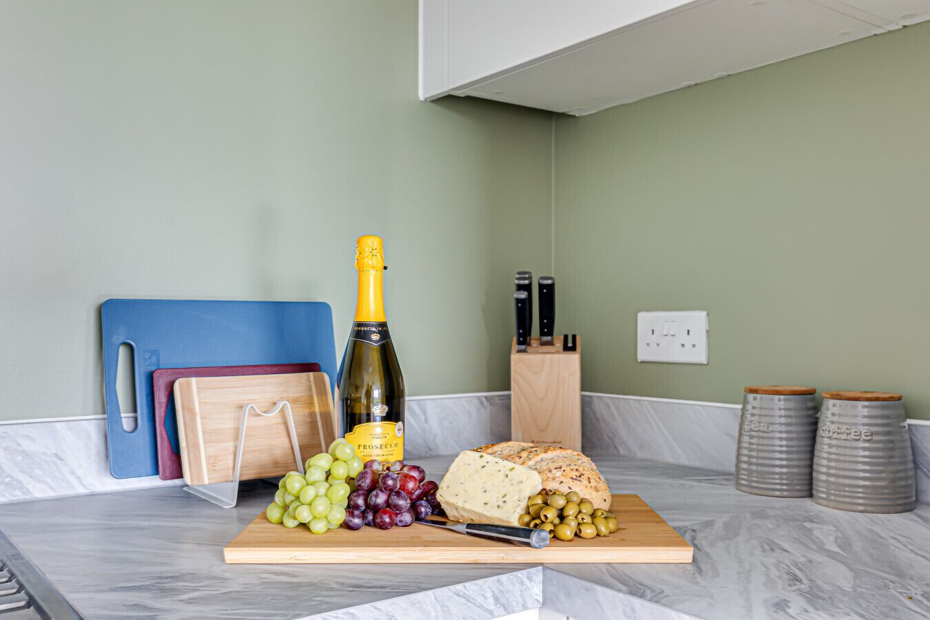 A kitchen counter with a cutting board holding a bottle of Prosecco, grapes, olives, and cheese.