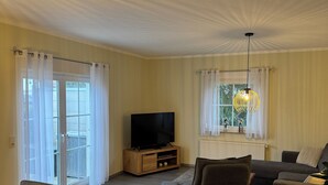 Ein Wohnzimmer mit Flachbildfernseher, einer Couch, einem Esstisch mit Stühlen und einem großen Fenster mit Vorhängen.