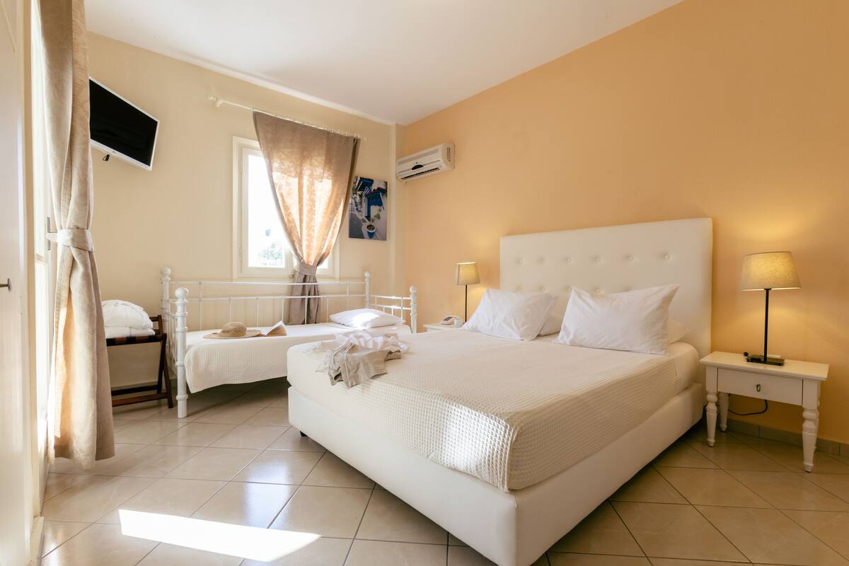 Une chambre avec un grand lit, un plus petit lit, une table de chevet avec une lampe, un téléviseur fixé au mur et une fenêtre avec des rideaux.