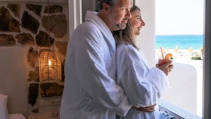 A couple in a white bathrobe standing by a window overlooking a beach. - Mikri Vigla Hotel Beach Resort (Naxos)