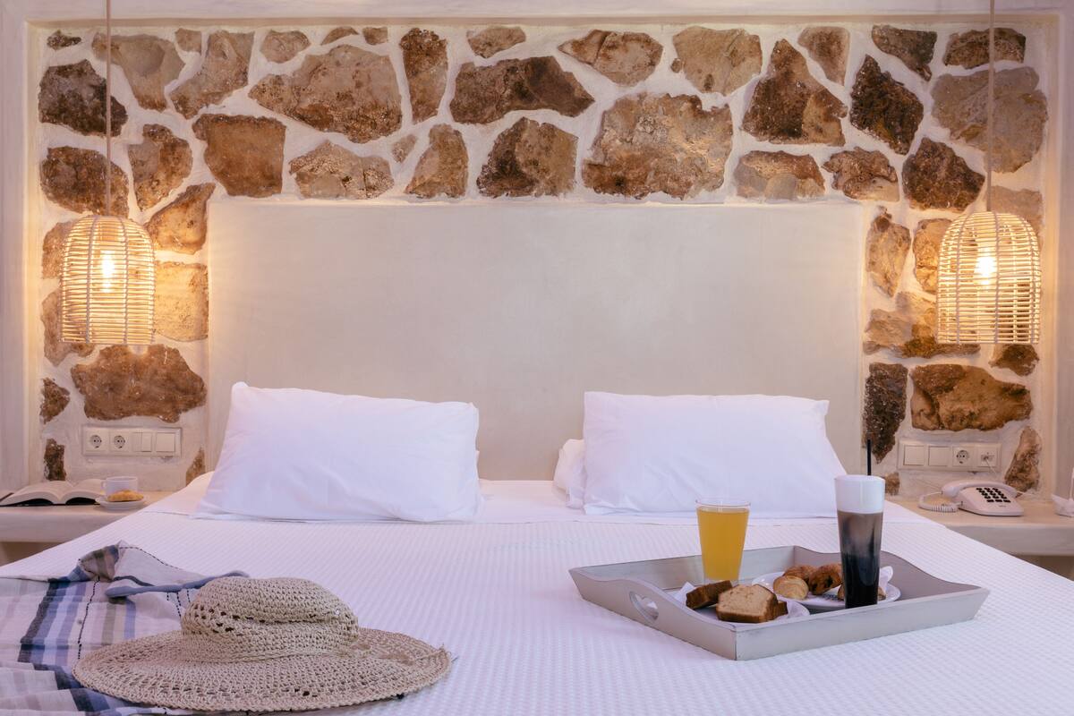 Une chambre d’hôtel avec un mur en pierre, deux lits blancs, un plateau avec des articles pour le petit-déjeuner et un chapeau de paille posé sur l’un des lits.