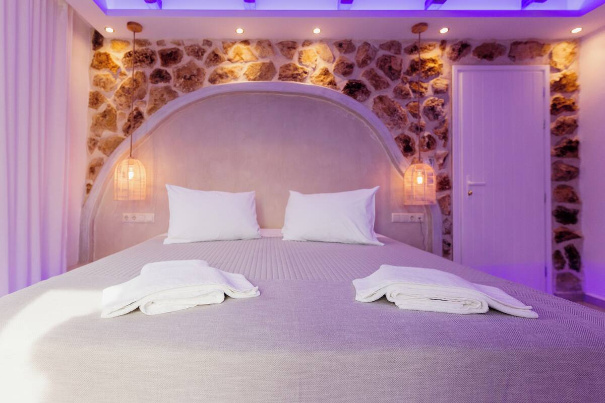 Une chambre avec un mur en pierre, un lit recouvert de draps blancs et deux lampes fixées au mur.