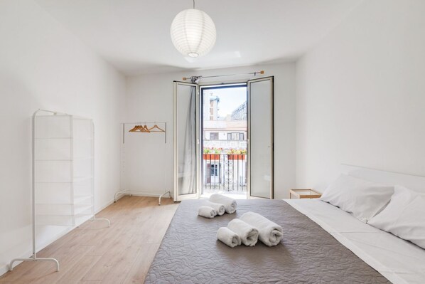 A hotel room with a bed, a hanging light fixture, a view through an open door, and a balcony with a railing.