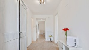 Un pasillo con una puerta blanca, un radiador, una mesita con mantel amarillo y un altavoz blanco sobre un soporte blanco.