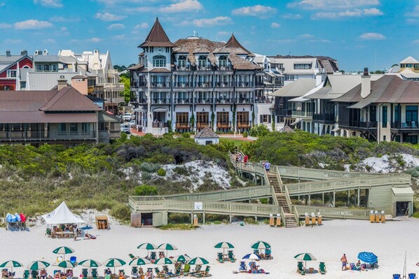 A beachfront hotel with a wooden boardwalk leading to the beach, multiple beach umbrellas, and a variety of building styles.