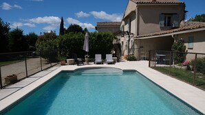Una piscina rectangular con borde de azulejos, rodeada por una valla. Hay sillas de descanso y una pérgola con sombrilla.