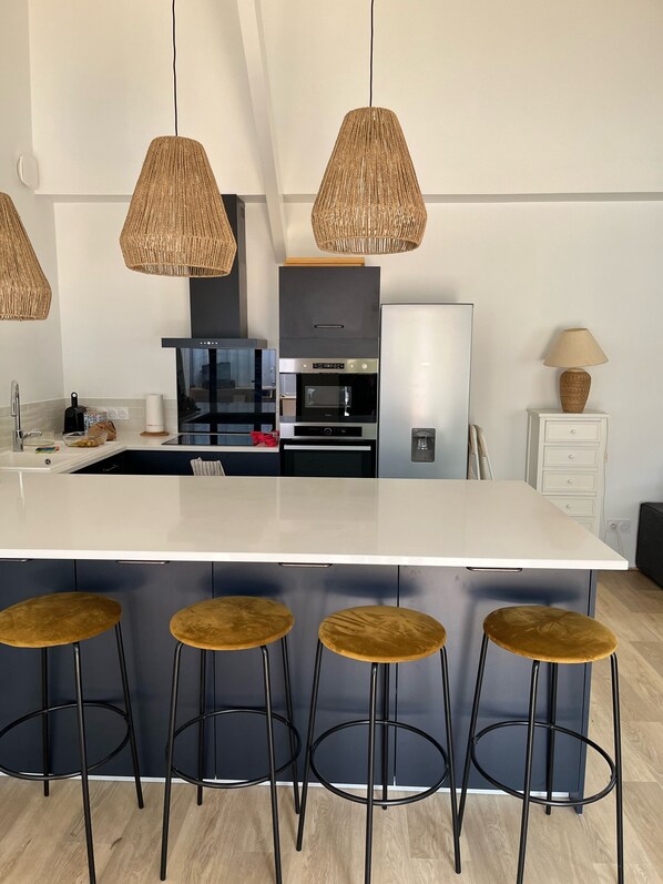 Una cocina moderna con una isla grande, taburetes negros, lámparas colgantes de mimbre y un armario blanco.