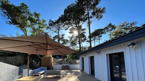 A wooden deck with outdoor seating, a large umbrella, and a building with a dark door. - Charming villa in Cap-Ferret (Lège-Cap-Ferret)