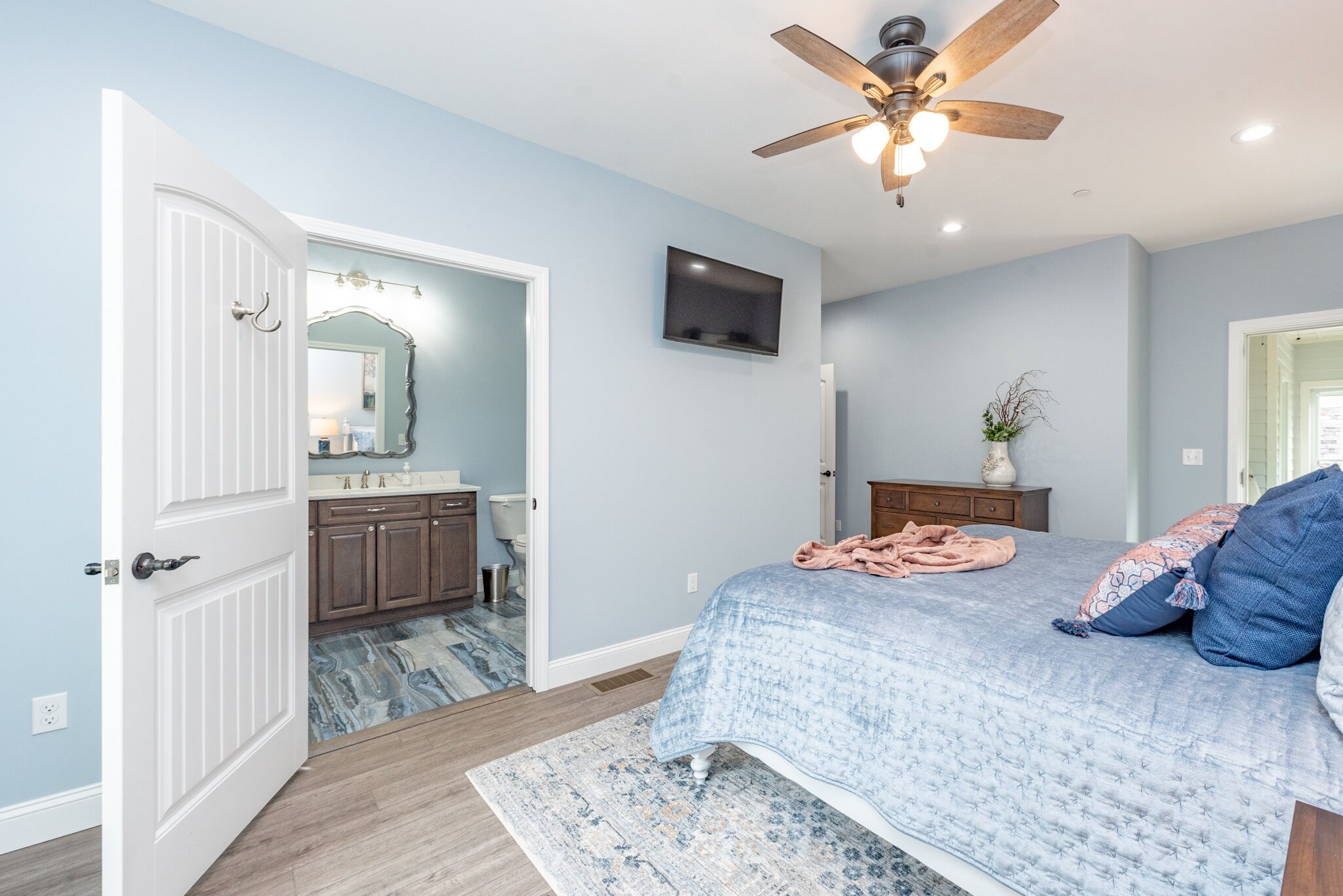 A bedroom with a bed, a ceiling fan, a TV mounted on the wall, and a bathroom visible through an open door.