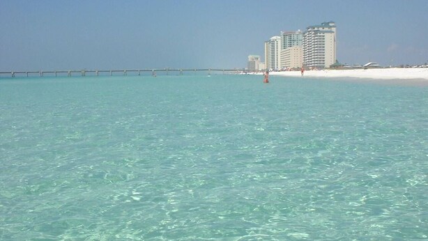 A beach with clear turquoise water, a pier extending into the sea, and several high-rise buildings in the background.