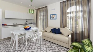 A modern living room with a sofa, a dining table with chairs, a kitchen area, and large windows with sheer curtains.