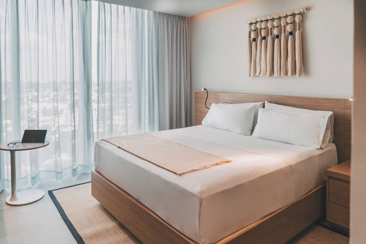 A modern hotel room with a large bed, a bedside table with a laptop, and a view of the city through sheer curtains.