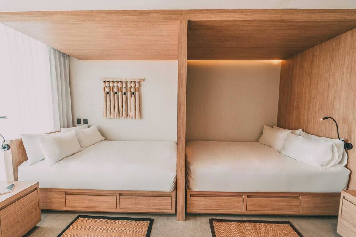 A modern hotel room with two beds, wooden headboards, a wall-mounted light fixture, and a woven rug.
