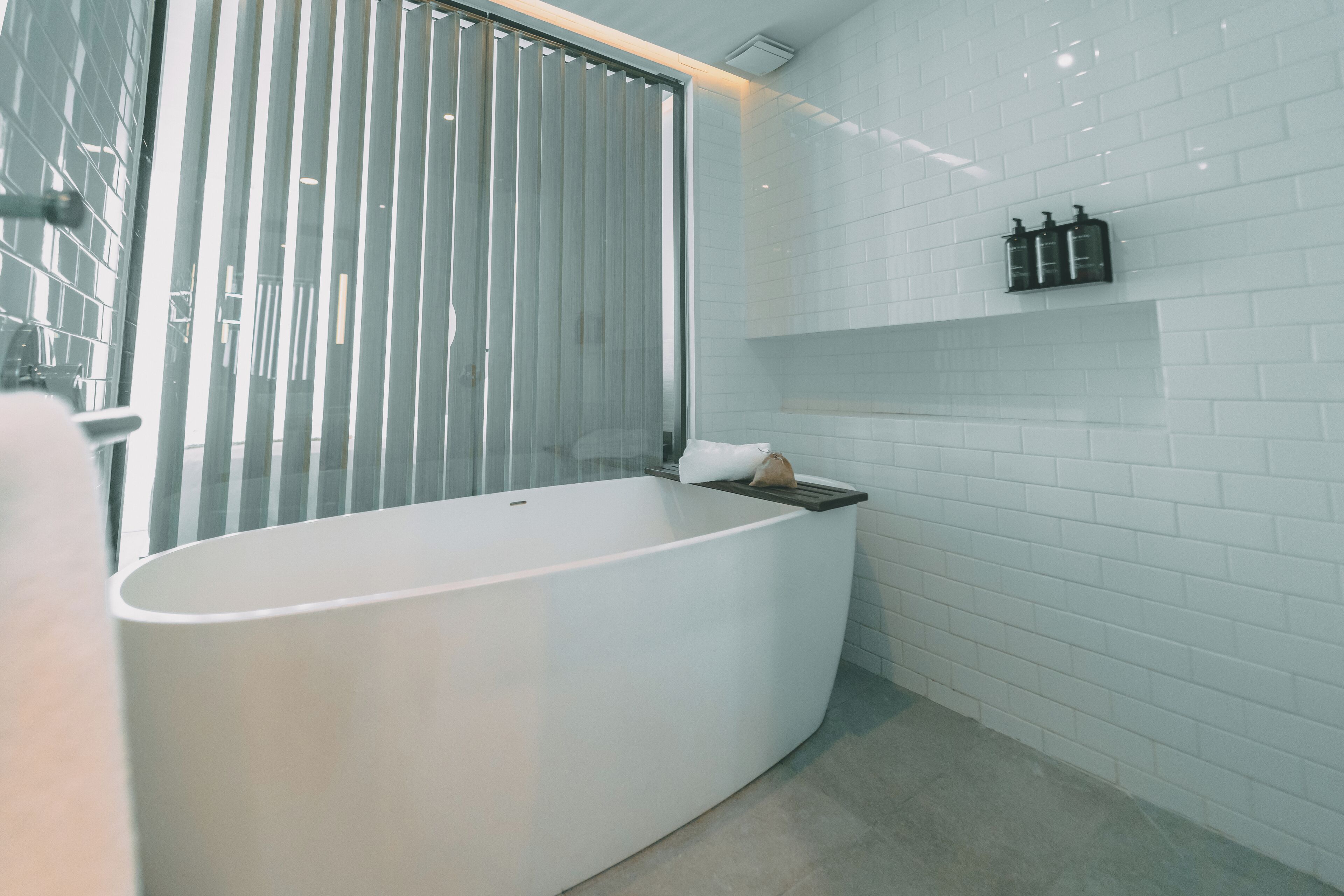 Un bagno moderno con una vasca freestanding, un box doccia in vetro e una parete rivestita di piastrelle bianche con uno scaffale per articoli da toilette.