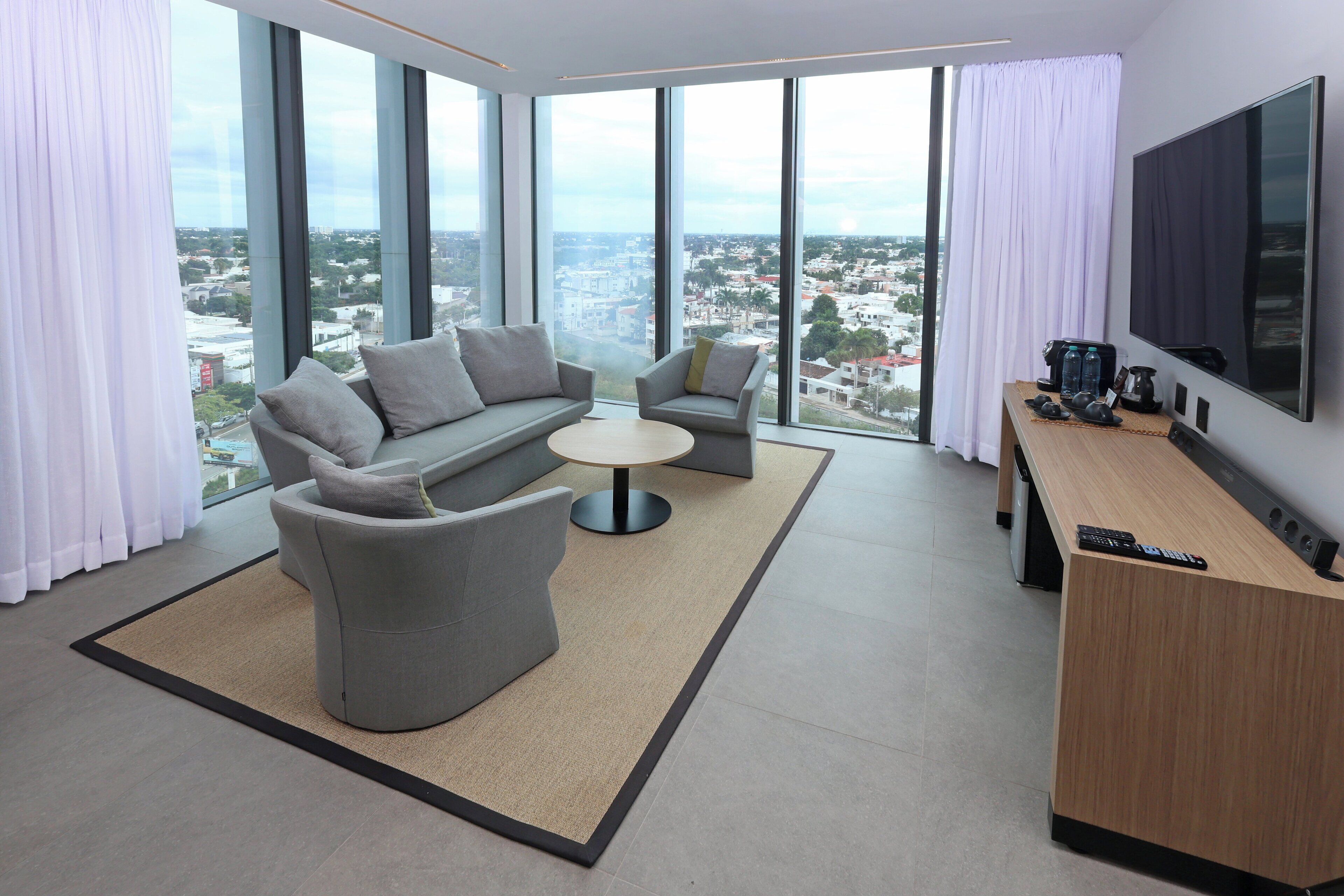 A modern hotel room with a sofa, armchair, and a small table. There is a TV mounted on the wall and a large window offering a city view.