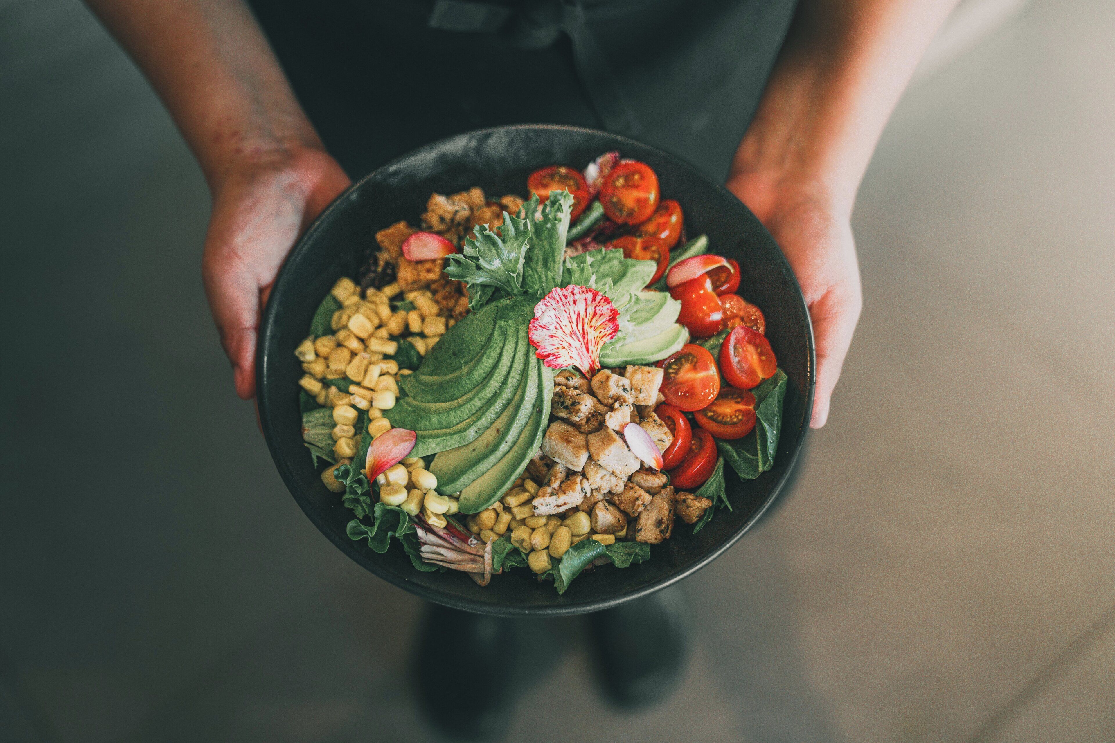 A person holding a bowl of salad with various vegetables and chicken.