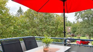 Un balcón exterior con mesa y sillas, un paraguas rojo y vistas a una zona residencial.
