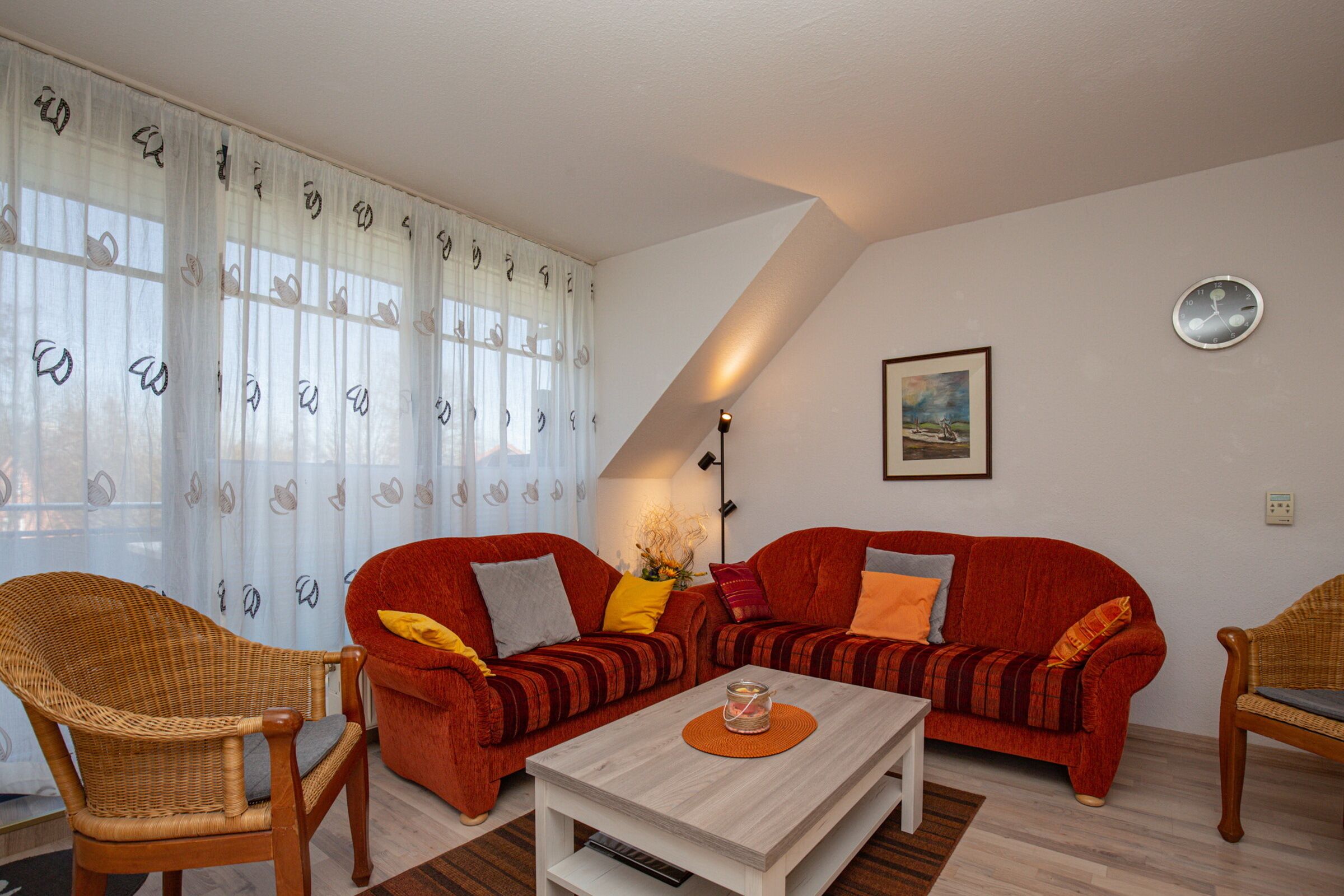Un salon avec deux canapés rouges, un fauteuil en osier, une table basse blanche et une grande fenêtre ornée de rideaux légers.