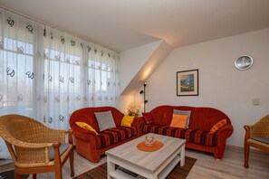Ein Wohnzimmer mit zwei roten Sofas, einem Korbsessel, einem weißen Couchtisch und einem großen Fenster mit durchsichtigen Vorhängen.