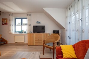 Ein gemütliches Wohnzimmer mit einer Couch, einem Korbstuhl, einem Fernseher und einer Küchenzeile mit Schränken und einem Fenster.
