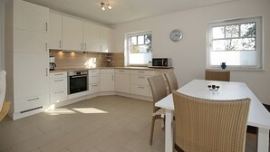 Une cuisine moderne avec des meubles blancs, un four encastré, un four à micro-ondes et un coin repas comprenant une table blanche et des chaises en osier.