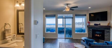A modern living room with a fireplace, a ceiling fan, and large windows offering a view of the outdoors.