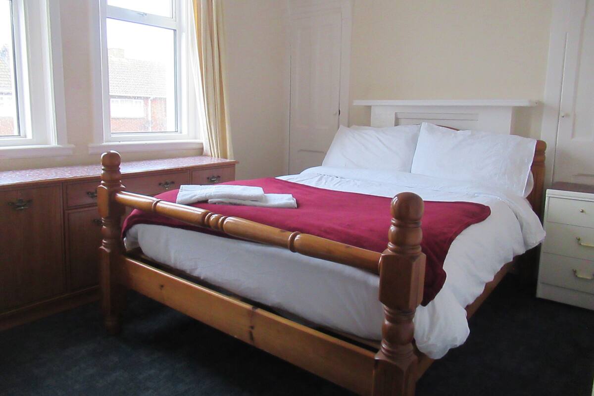 Uma cama bem arrumada com estrutura de madeira, roupa de cama branca e manta vermelha. Há uma mesinha de cabeceira de madeira com gaveta e uma janela com cortinas à esquerda.