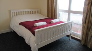 A single white bed with a red blanket and two rolled towels, a white headboard, and a window with curtains. - Lennard House (Folkestone)